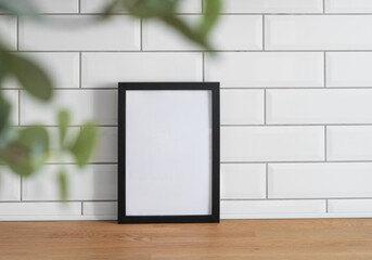 Wall Mural - A black frame with a blank canvas against a white tile wall and on a wooden tabletop with blurred green foliage.