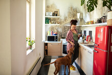 Young woman feeding dog while using smartphone in home kitchen