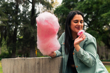 Wall Mural - beautiful latin woman sitting, smiling and eating her cotton candy, enjoying the day, in an outdoor park