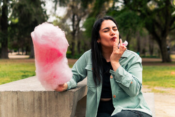 Wall Mural - beautiful latin woman sitting, smiling and eating her cotton candy, enjoying the day, in an outdoor park