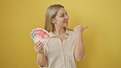 Sticker - Joyful young woman flashing a thumbs up, grinning with banknotes of israel shekel in hand, on isolated yellow wall