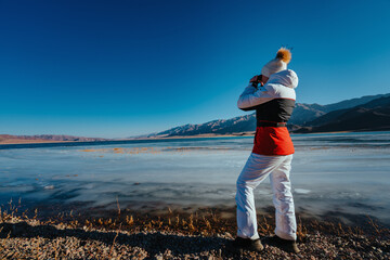 Wall Mural - Young woman tourist standing on shore of lake and looking through binoculars in winter