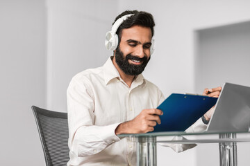 Aufkleber - Happy indian man in headphones using tablet and laptop at modern office desk