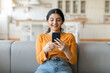 © Prostock-studio - Beautiful young indian woman using her smartphone while sitting on couch