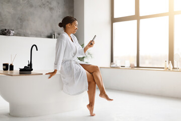 Wall Mural - Black woman in white bathrobe laughing while texting on her smartphone