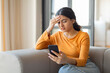 © Prostock-studio - Indian woman sitting on couch at home and looking at smartphone screen