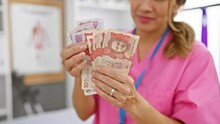 A Hispanic Woman In Pink Scrubs Counts Colombian Pesos In A Hospital Room.
