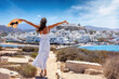 A happy tourist woman in a white summer dress looks at the whitewashed houses of the town of Naxos island, Cyclades, Greece