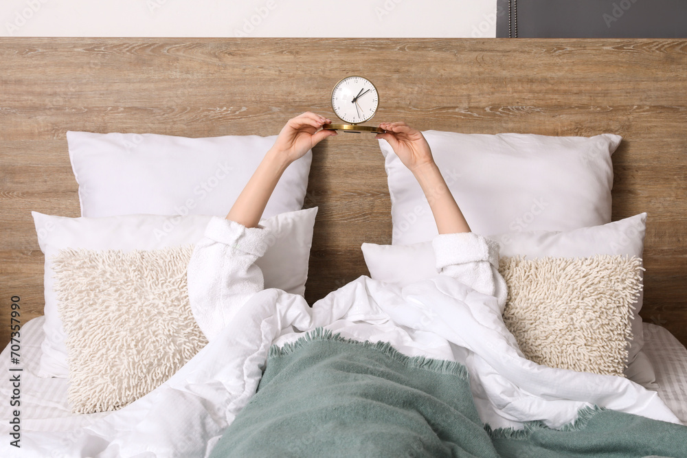 Woman in pajamas holding alarm clock while lying on bed