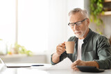 Fototapeta  - Concentrated elderly male in glasses drinking coffee while reviewing documents at home