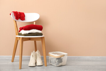 Stack of folded sweaters with gloves on chair and boots near beige wall