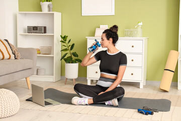 Poster - Sporty young African-American woman drinking water after training in living room