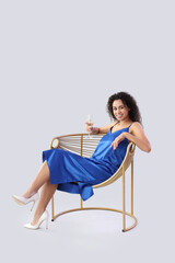 Young African-American woman with glass of champagne sitting in armchair on light background