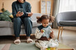 Full shot of African American girl with two ponytails playing with toys on living room floor while unrecognizable mother on sofa holding smartphone and thermometer