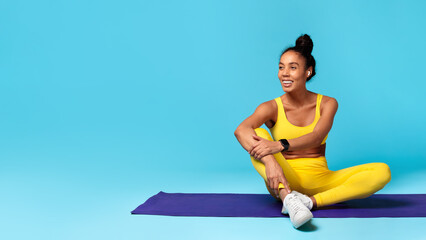 Wall Mural - athletic african american woman sitting on yoga mat, blue background