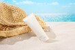 Close-up of an unlabeled tube of sunscreen leaning against a straw hat on white sand and blue sea water. Exquisite mockup for advertising. Blank labels for branding.
