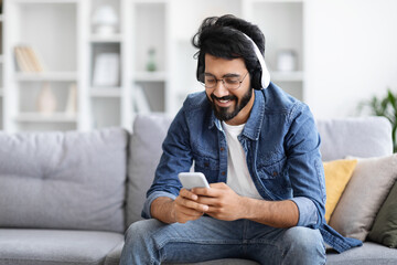 Wall Mural - Relaxed indian man listening music on smartphone with wireless headphones at home