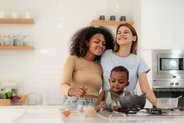 Wall Mural - Multiracial lesbian couple cooking healthy food in kitchen for their biracial boy. Gay women standing at counter enjoy preparing lunch at home with child kid. Young adult LGBT family eating together