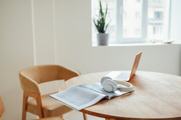 Laptop, headphones and magazine on a round wooden table next to a comfortable beige armchair.