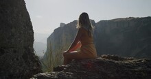 A Young Woman Sitting On The Rocks Of Meteora. Sweeping Slow Motion Gimbal Shot, Hair Blowing In The Wind.