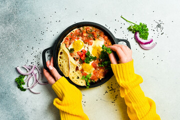 Wall Mural - Hands hold a frying pan with Shakshuka. Poached eggs in a spicy tomato pepper sauce. Traditional Jewish scrambled eggs. Top view, flat lay.