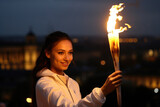 Fototapeta  - Young woman holding a torch with a bright flame at dusk, city lights blurred in the background.