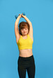 Teen girl with down syndrome in sportswear training, lifting dumbbells against blue studio background. Concept of acceptance, care, inclusion, health, diversity, emotions, equality