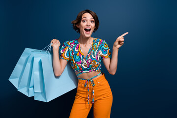 Wall Mural - Photo of lovely young lady shopping bags point empty space dressed stylish flared pants garment isolated on dark blue color background