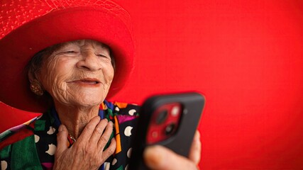 Wall Mural - Funny fisheye view of old woman with no teeth having video chat on phone with family and friends isolated on red background.