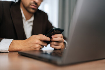 Close-up cropped shot of unrecognizable businessman in suit scrolling on smartphone screen reading content while working on laptop sitting office desk. Male entrepreneur research, web scrolling, email