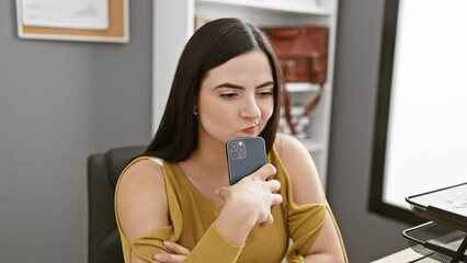 Wall Mural - Thoughtful hispanic woman with smartphone pondering in office setting