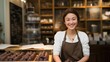 A joyful young woman wearing a white shirt and brown apron stands proudly in her chocolate shop, showcasing an array of gourmet treats
