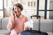 Cropped old elderly woman grandmother traveler talking on the phone, calling ordering taxi to airport, train station with her luggage baggage suitcases bags at home with airline tickets