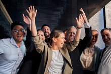 Diverse Multiracial Cheerful Group Of Excited Business People Standing In Office Hallway Having Fun. Joyful Colleagues Celebrating Success By Raising Hands At Same Time And Smiling Happy Together