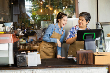 Young barista female hiring senior, elderly, 60s pensioner worker working in cafe bakery small business shop, two waitresses women standing behind coffee counter using technology cash register machine