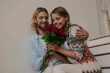 Romantic young man giving a bouquet of roses to his happy girlfriend while sitting on stairs at home