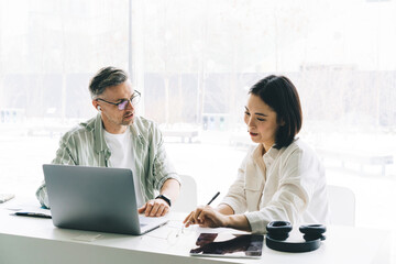 Fototapete - Male employee in optical eyeglasses communicate with Asian female colleague during cooperation process at table desktop with laptop technology, professional entrepreneurs talking about business