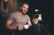 Portrait of happy Caucasian man with coffee cup and modern smartphone technology smiling at camera during free time, carefree bearded male customer with mobile gadget posing during break in cafe