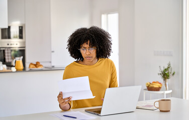 Young worried stressed anxious African American woman checking financial paper calculating banking loan or household payment using laptop computer paying bills online sitting at home table in kitchen.