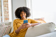 © insta_photos - Smiling young African woman using cell phone relaxing on couch at home. Happy lady sitting on sofa looking at smartphone holding mobile cell phone in hands texting buying online in ecommerce shop.