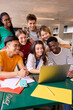 © CarlosBarquero - Vertical large group of young multiracial students gathered in classroom smiling and having fun watching something funny on laptop. Generation z friends using and looking screen computer inside