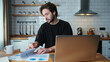 © mahirkart - Young curly hair man working at home in the kitchen working with his laptop, doing research with papers. Busy freelancer working on modern tech notebook device