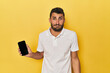 © Asier - Young Hispanic man shows phone screen shrugs shoulders and open eyes confused.