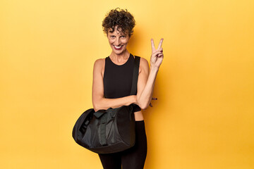 Wall Mural - Sportswoman with gym backpack on yellow studio showing number two with fingers.