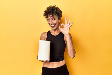 Wall Mural - MIddle aged athlete woman holding protein supplement on yellow cheerful and confident showing ok gesture.