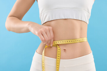 Slim woman measuring waist with tape on light blue background, closeup. Weight loss