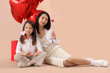 Wall Mural - Young Asian woman and her little daughter holding paper hearts and air balloons on brown background. Valentine's day celebration