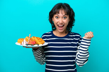 Wall Mural - Young Argentinian woman holding waffles isolated on blue background celebrating a victory in winner position