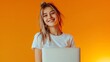 Enjoying student life. Portrait of young smiling girl in white t-shirt with laptop isolated on orange color background in neon light. Concept of beauty, art, fashion, education   