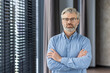 © Liubomir - Confident mature businessman with glasses standing with arms crossed in modern office setting, conveying leadership and professionalism.